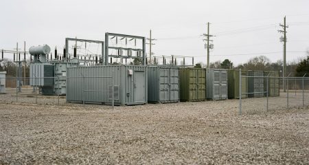 A row of container-sized battery storage units on a fenced site beside a substation