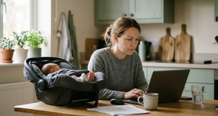 A parent working at a laptop in a home kitchen with a baby carrier on the table beside them